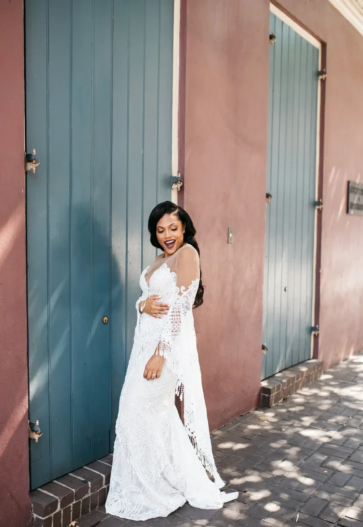 A woman in a white lace dress standing next to a pink wall with teal doors, smiling and looking down.