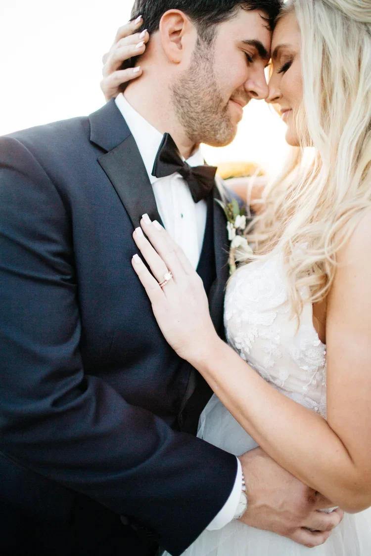 A bride and groom in wedding attire smiling and holding each other closely, with foreheads touching, outdoors during sunset.