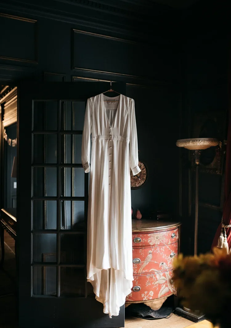 A white long dress hanging on a black hanger in a dark room with vintage decor and a small curved red chest of drawers.