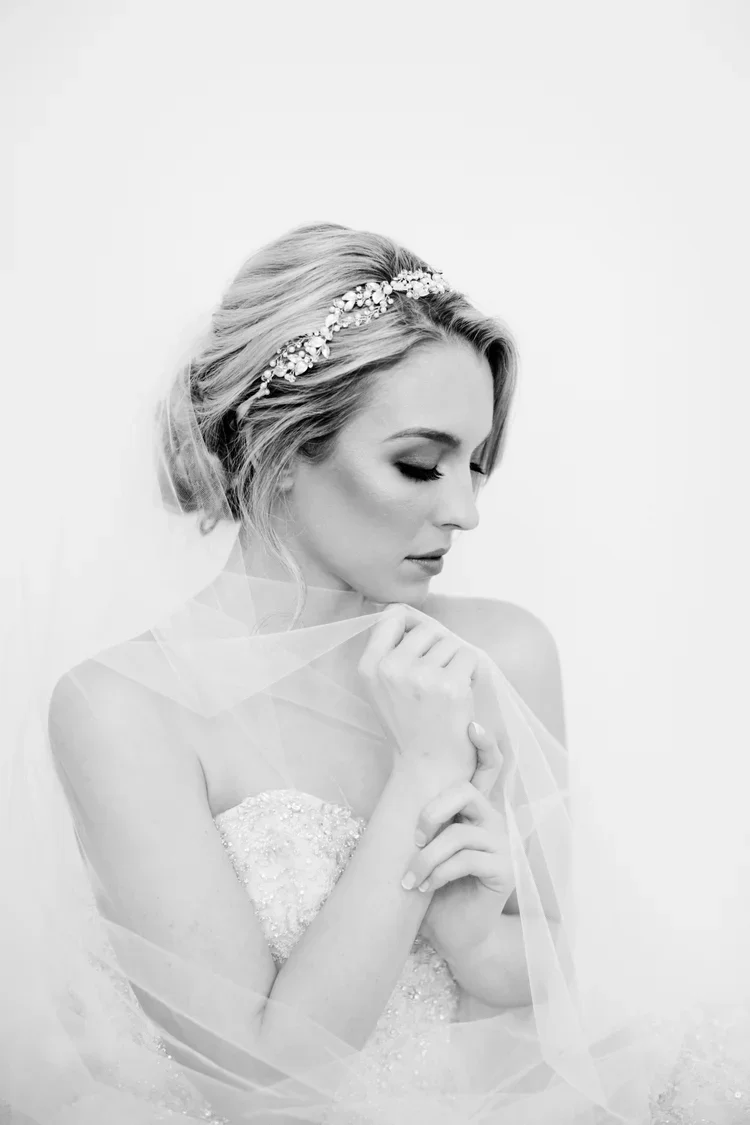 Black and white portrait of a bride with her eyes closed, wearing a tiara and a strapless wedding dress, holding a sheer veil close to her chest.