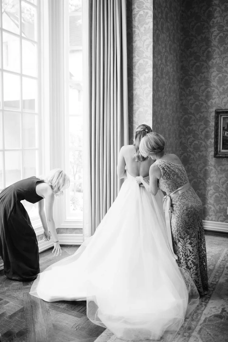 Three women preparing a bride in a wedding dress in a well-lit room with large windows, curtains, and patterned wallpaper.