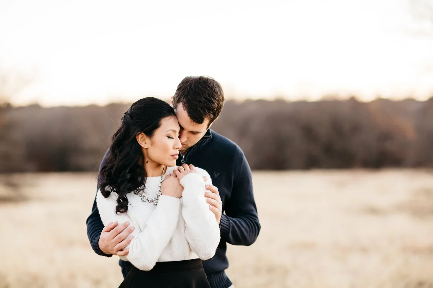 A couple standing close together outdoors during sunset, with their foreheads touching in an intimate moment. The woman has long dark hair and is wearing a white sweater and a necklace, while the man has short brown hair and is dressed in a dark jack