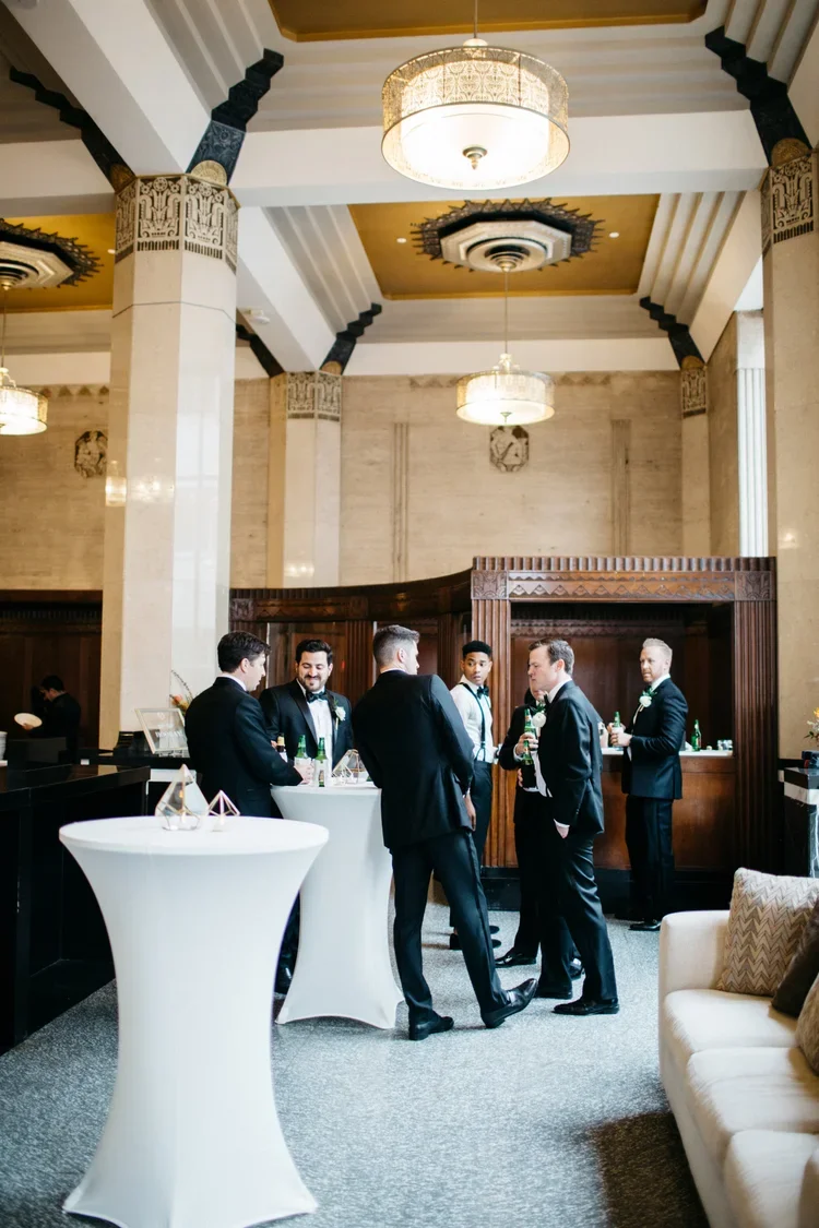 Group of men in tuxedos standing and talking at a formal event in an elegant, decorated room with tall columns, chandeliers, and a bar area.