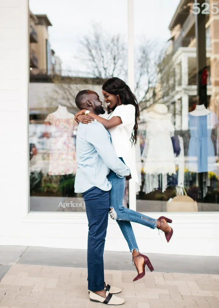 A couple hugging outside a clothing store, with the woman lifting her legs and smiling at the man. The store window displays dresses and mannequins.