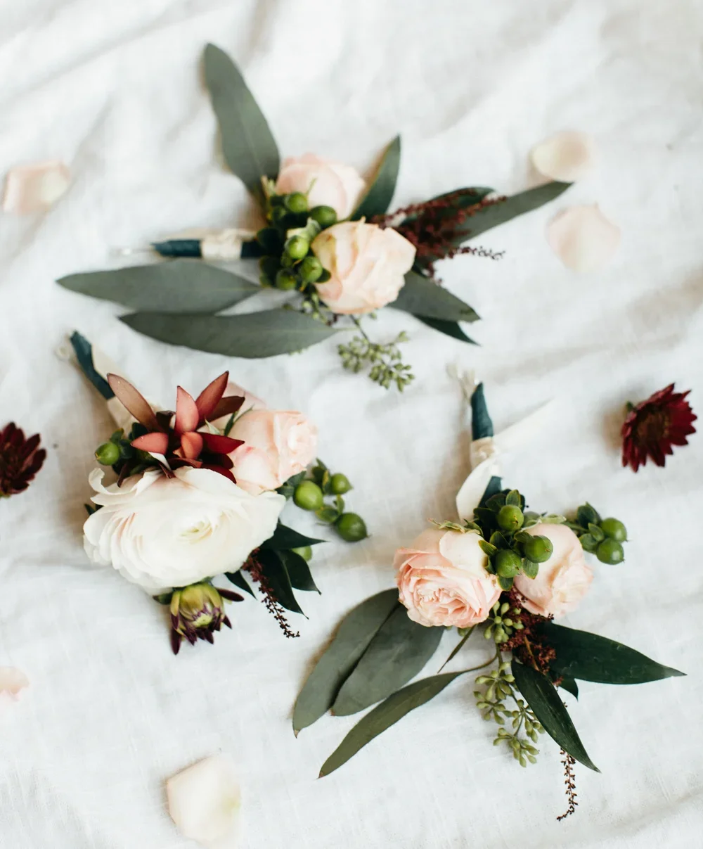 Three boutonnières with pale pink and white roses, green berries, eucalyptus leaves, and small dark red flowers, arranged on a white fabric with scattered rose petals.