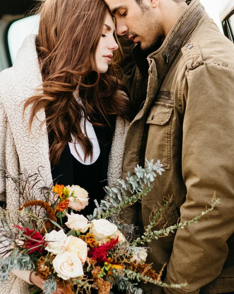 A couple stands close with their foreheads touching, holding a bouquet of flowers, outdoors.