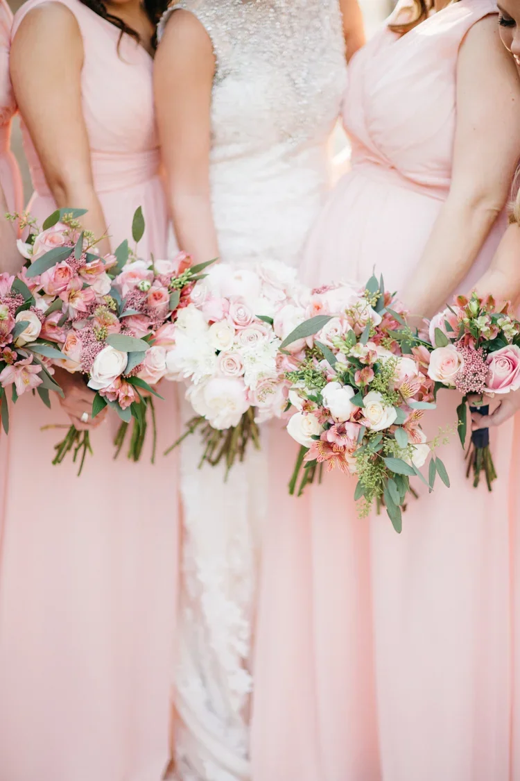 A bride in a wedding dress standing with four bridesmaids dressed in pink, each holding a colorful bouquet of pink and white flowers with greenery.