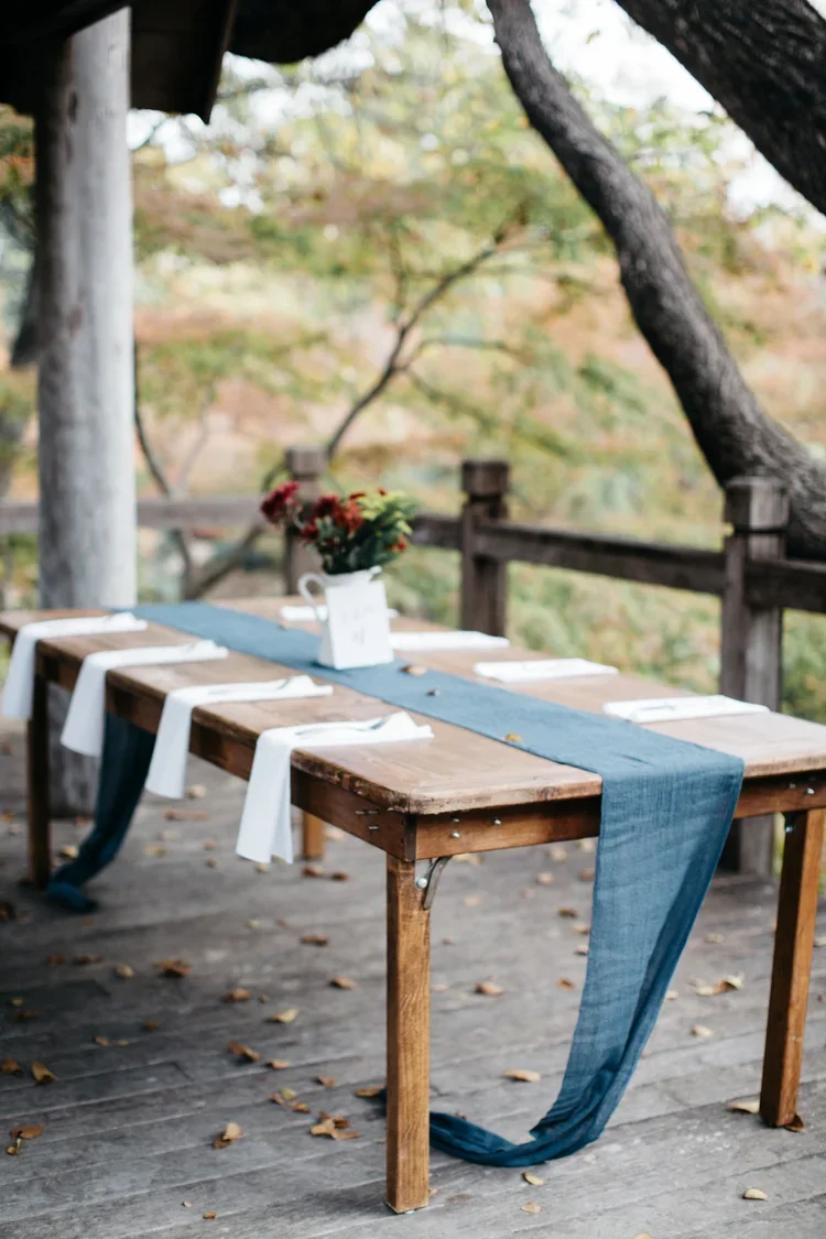 A rustic outdoor dining table set for a meal, decorated with a blue table runner, white napkins, and a flower arrangement in a white pitcher, on a wooden deck surrounded by trees.