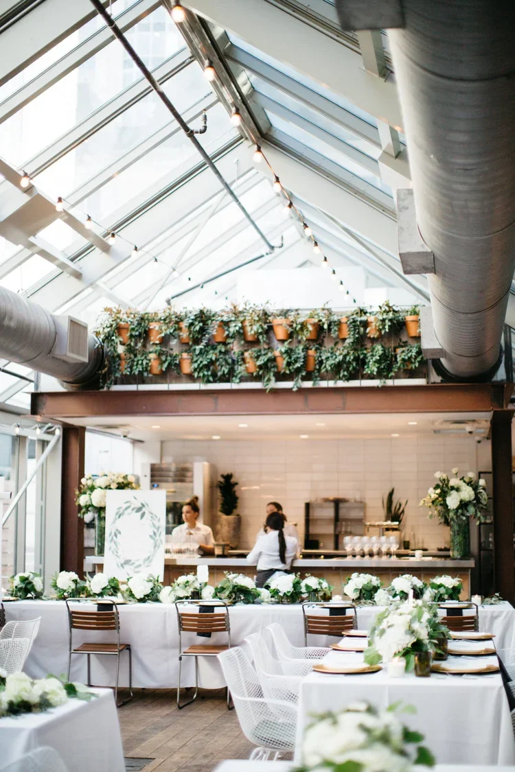 Indoor event space with a glass ceiling, decorated with white flowers and green foliage, set up for a wedding or celebration.