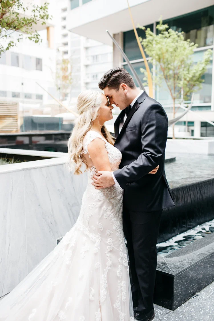 A bride and groom stand close together with foreheads touching, outdoors in an urban setting.