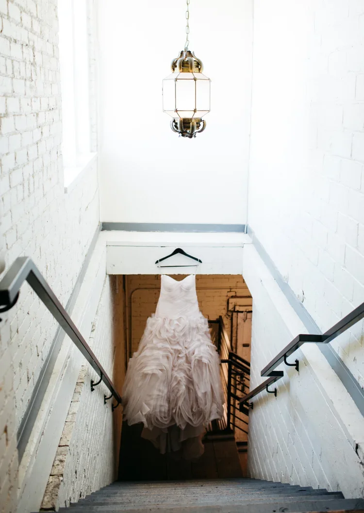 Wedding dress hanging on a hanger in a stairwell with white brick walls and a vintage chandelier hanging from the ceiling.