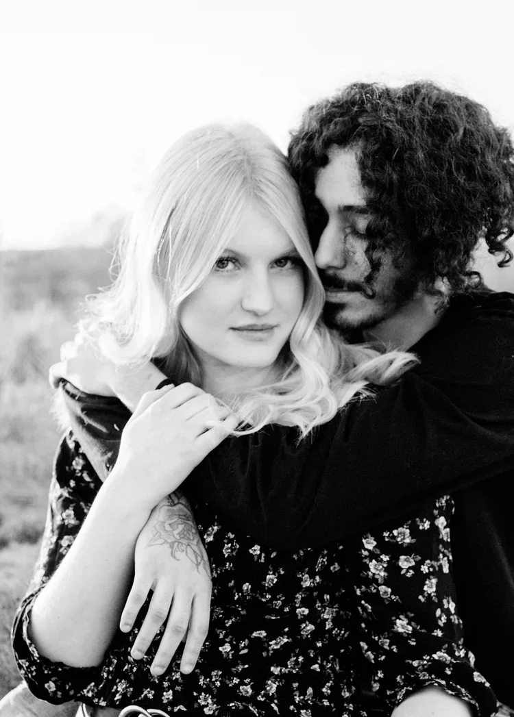 Black and white photo of a young couple embracing outdoors, woman with long blonde hair and tattoo on arm, man with curly hair, both facing the camera with the man’s face close to the woman’s.