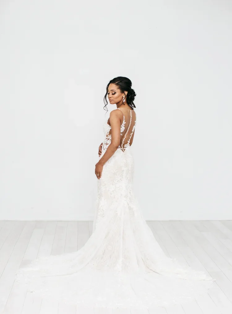 A woman in a fitted white wedding dress with lace details and an open back, standing against a plain white wall and floor.