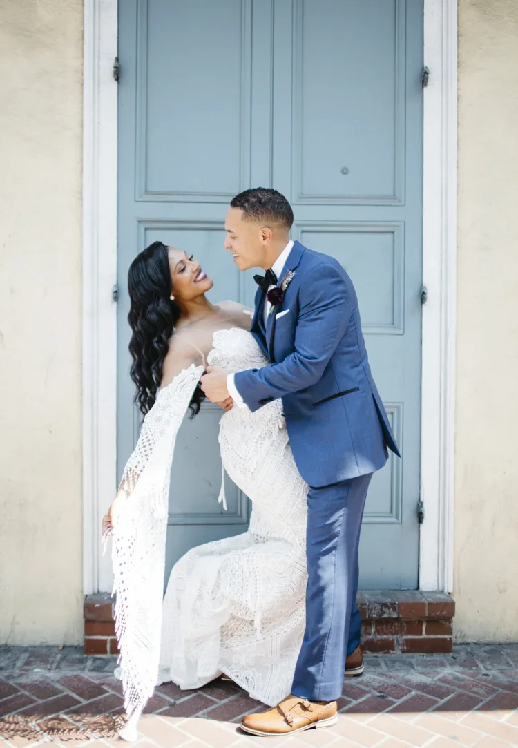 A newlywed couple smiling at each other in front of a blue door, with the bride dressed in a white lace wedding gown and the groom in a blue suit.