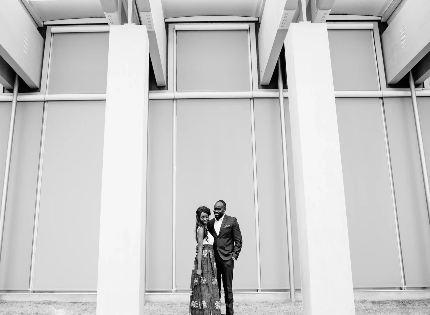 Black and white photo of a couple standing close, smiling, in front of a modern architectural building with large vertical panels.