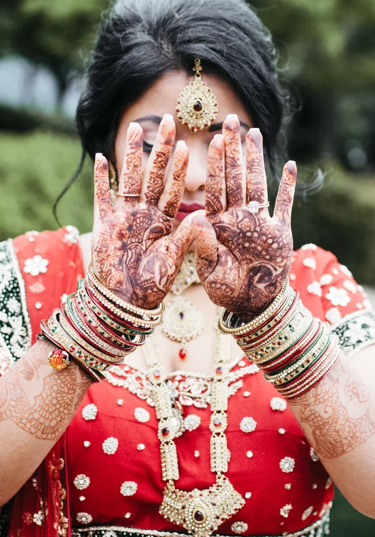 A woman dressed in traditional Indian bridal attire, showing her henna-decorated hands, jewelry, and earrings, with a garden in the background.