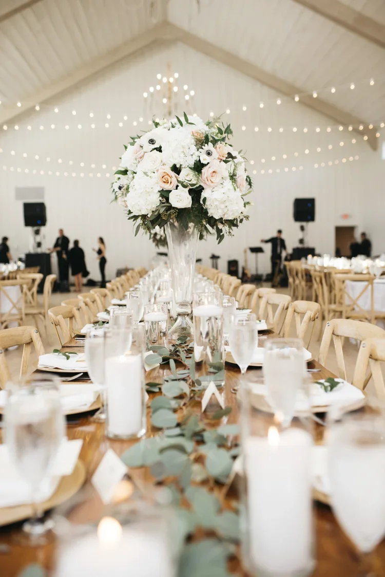 A large wedding reception hall with a long wooden table decorated with a green eucalyptus runner, candles, and place settings. A tall floral centerpiece with white and light pink roses and white hydrangeas is in the middle. String lights hang from th