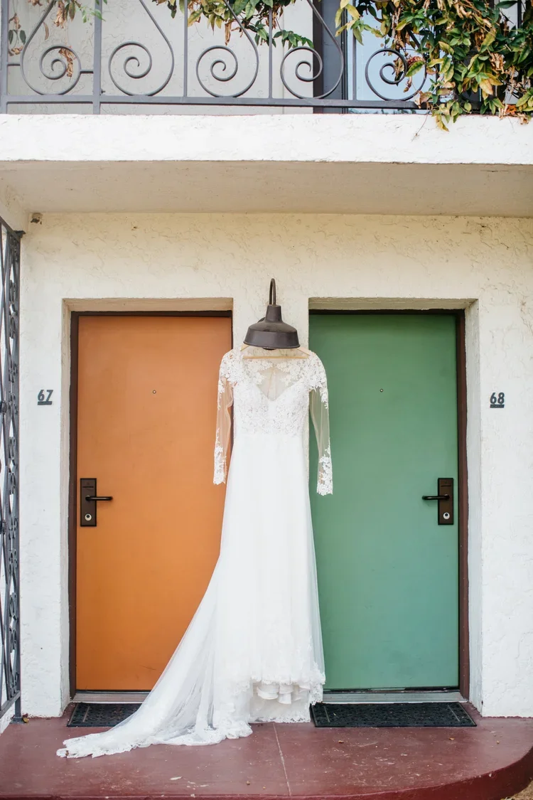 A wedding dress hanging in front of two apartment doors, one orange and one green, with a railing and greenery above.
