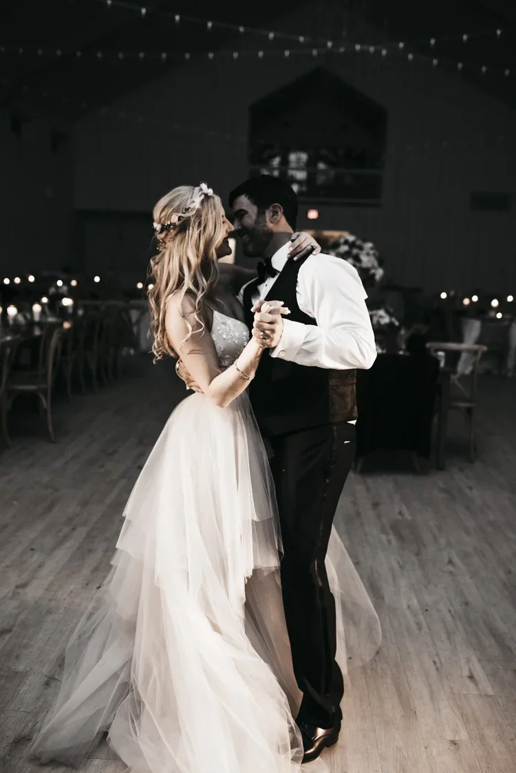 A bride and groom sharing a dance at their wedding reception in a dimly lit venue with string lights overhead and decorated tables in the background.