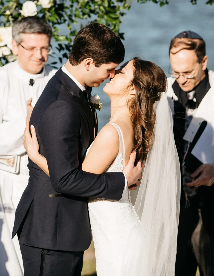 Bride and groom sharing a kiss during their wedding ceremony outdoors by the water, with two officiants smiling in the background.