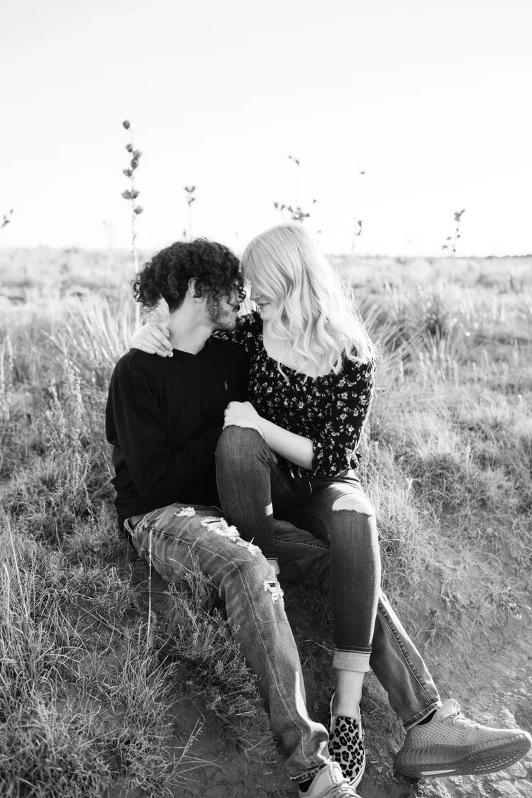 A black and white photo of a young couple sitting on the grass outdoors, close together, with their foreheads touching and smiling. The man has curly hair and is wearing a dark long-sleeve shirt and ripped jeans. The woman has long wavy blonde hair a