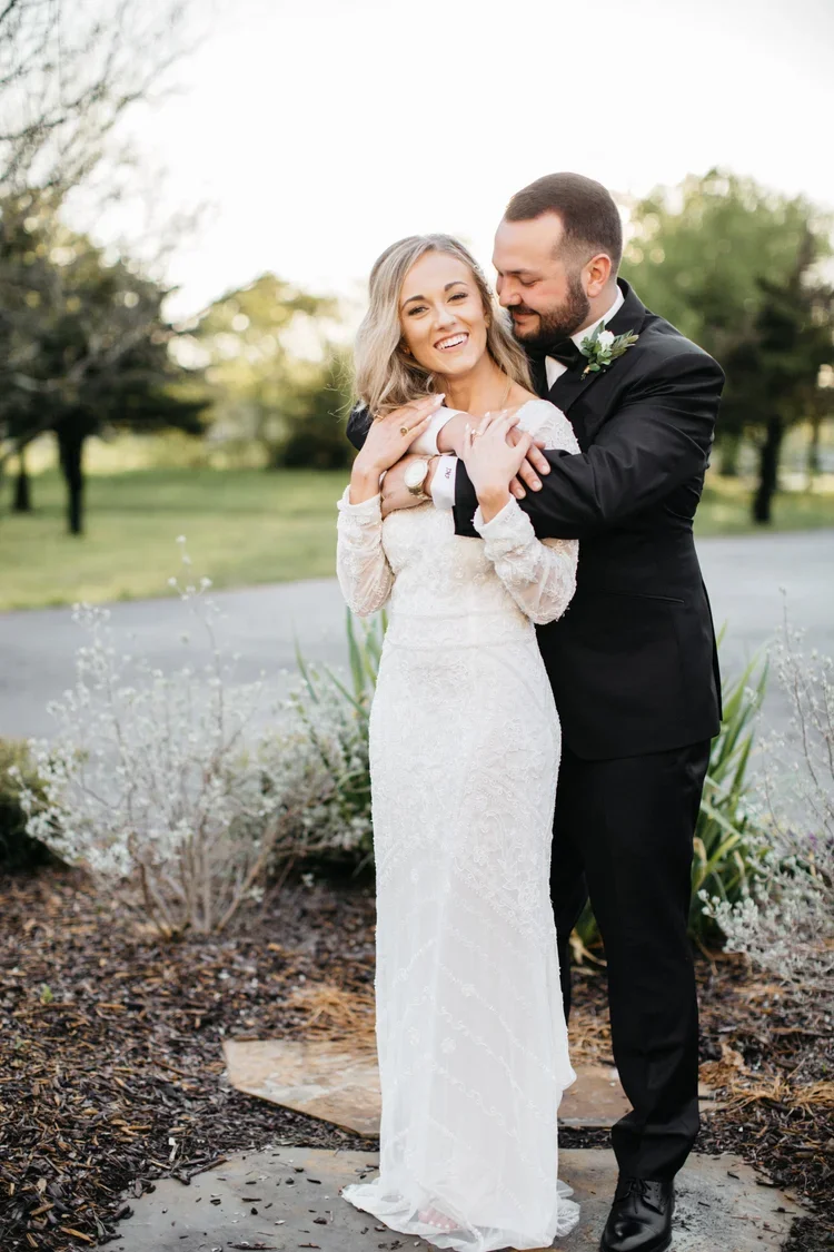 A newlywed couple in wedding attire hugging outdoors in a park or garden.