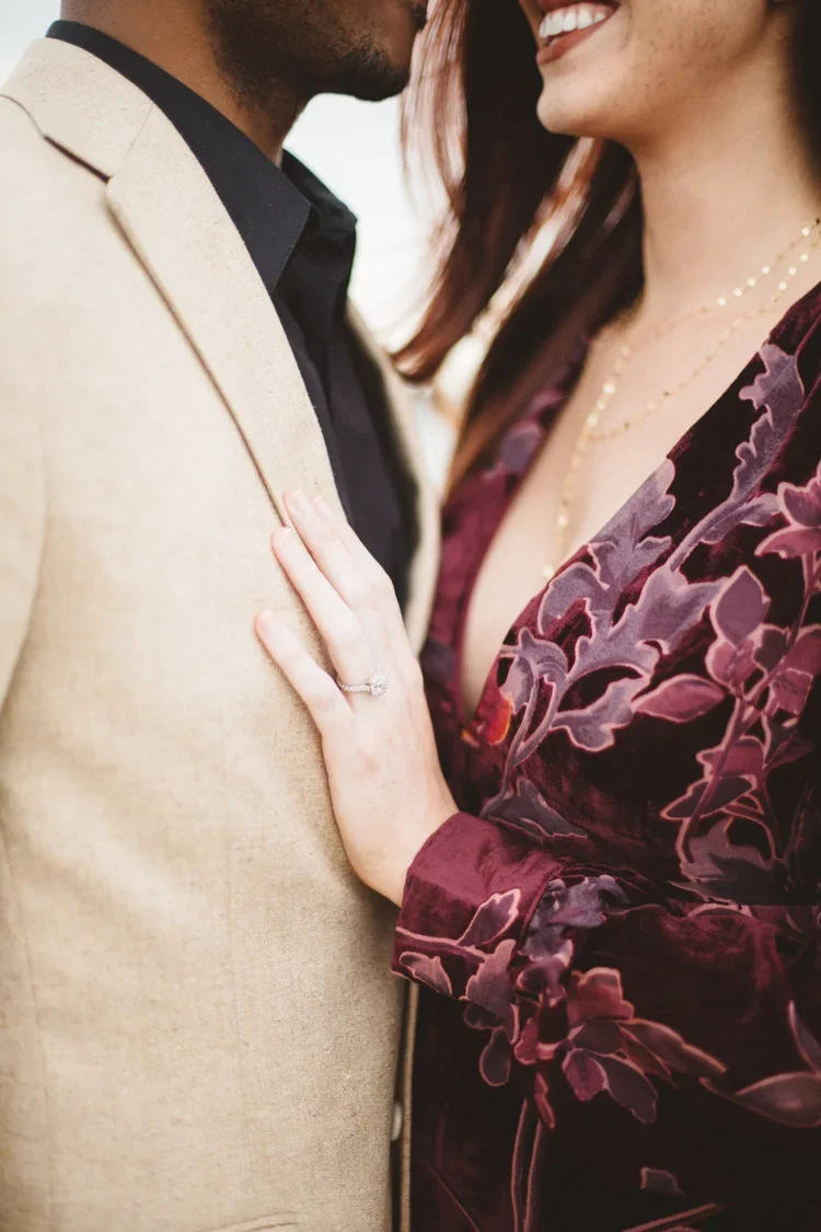 A close-up of a couple smiling and touching each other, with a focus on the woman's hand displaying an engagement ring.