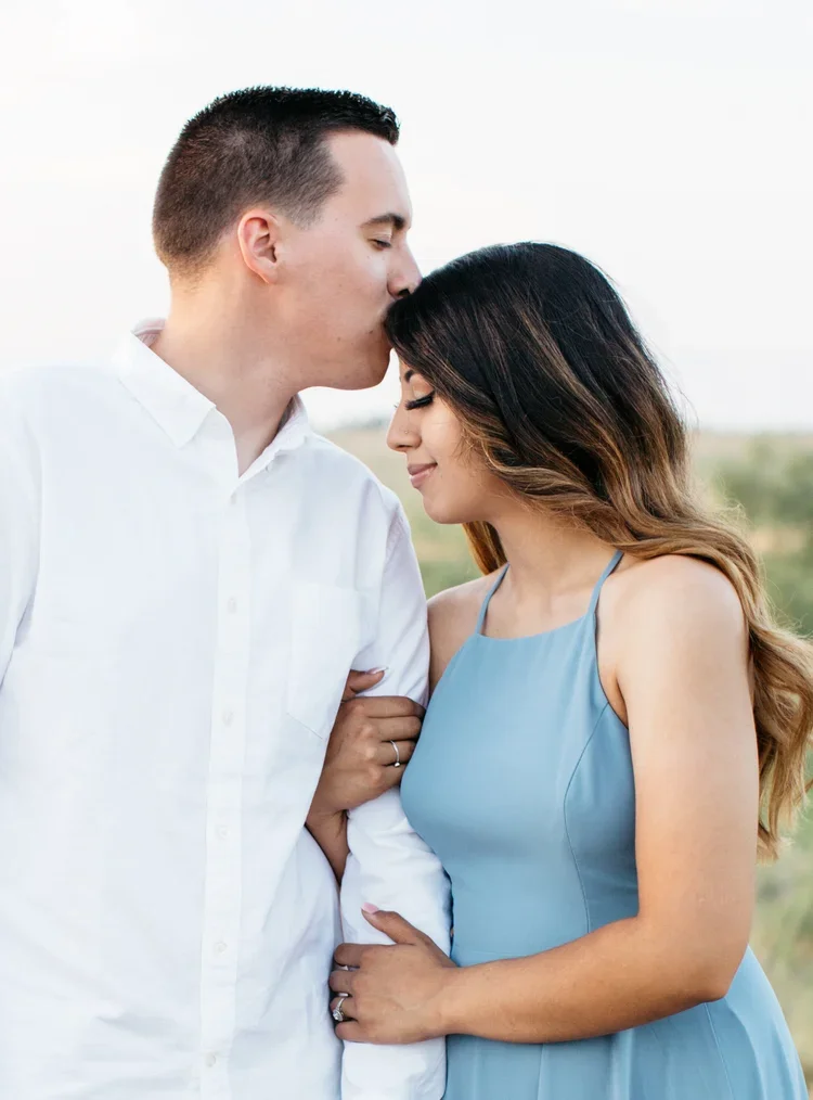 A man and woman sharing a tender moment outdoors, with the man kissing the woman's forehead and both smiling softly. The woman is wearing a light blue dress and the man is dressed in a white shirt.