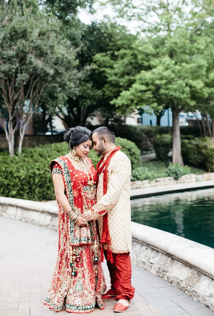 A couple dressed in traditional Indian wedding attire standing close together by a pond, with trees and greenery in the background.