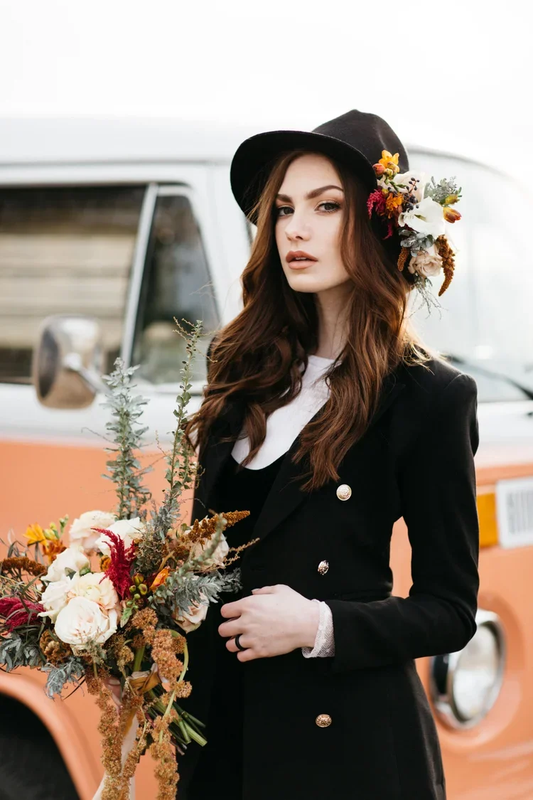 A woman with long brown hair wearing a black hat decorated with flowers, a black blazer with gold buttons, and holding a bouquet of flowers, standing in front of a vintage van.