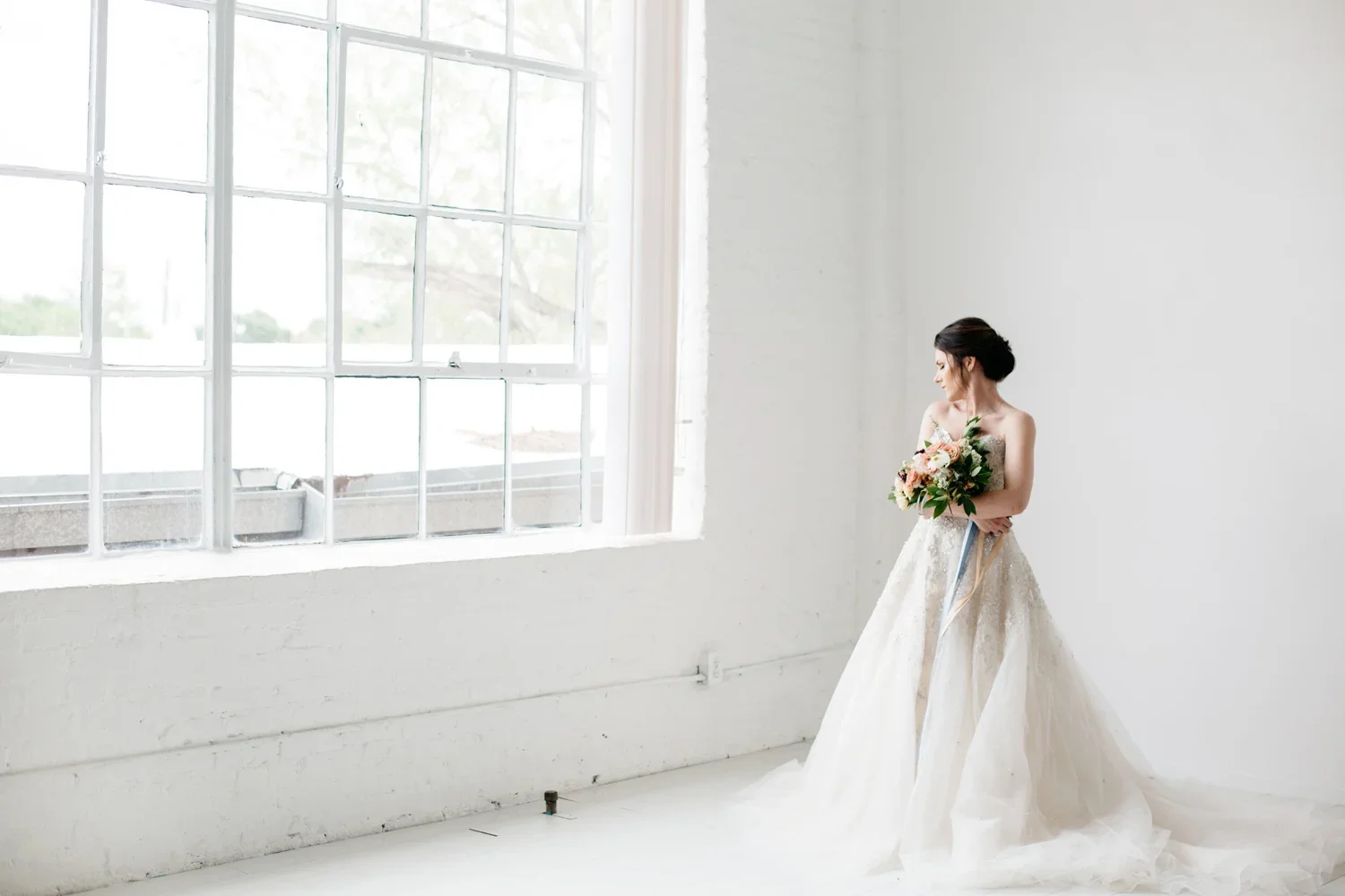 A bride in a wedding gown holding a bouquet of flowers, standing in a bright, minimalistic room with a large window.