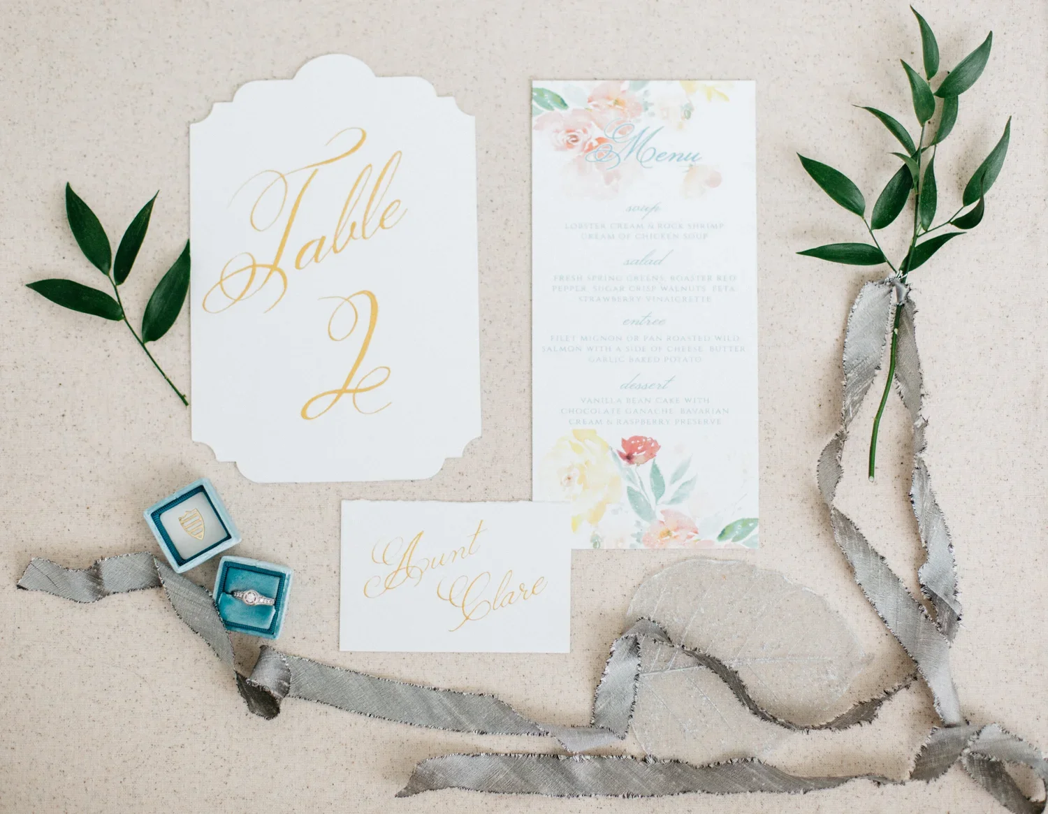 Wedding invitation with a floral menu, a table sign, and place cards for the bride and groom, decorated with green leaves and silver ribbon, on a beige surface.