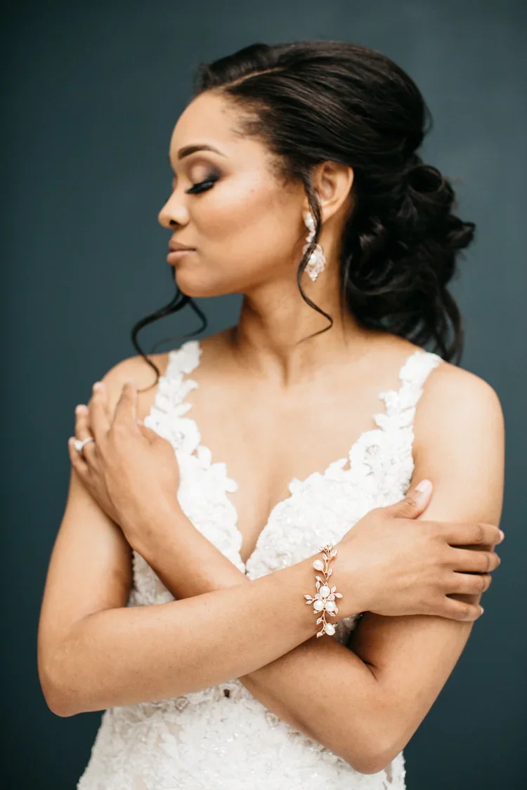 A woman in a white lace dress with floral details, wearing earrings and a bracelet, with dark hair styled in loose waves, standing with her arms crossed and eyes closed against a dark backdrop.