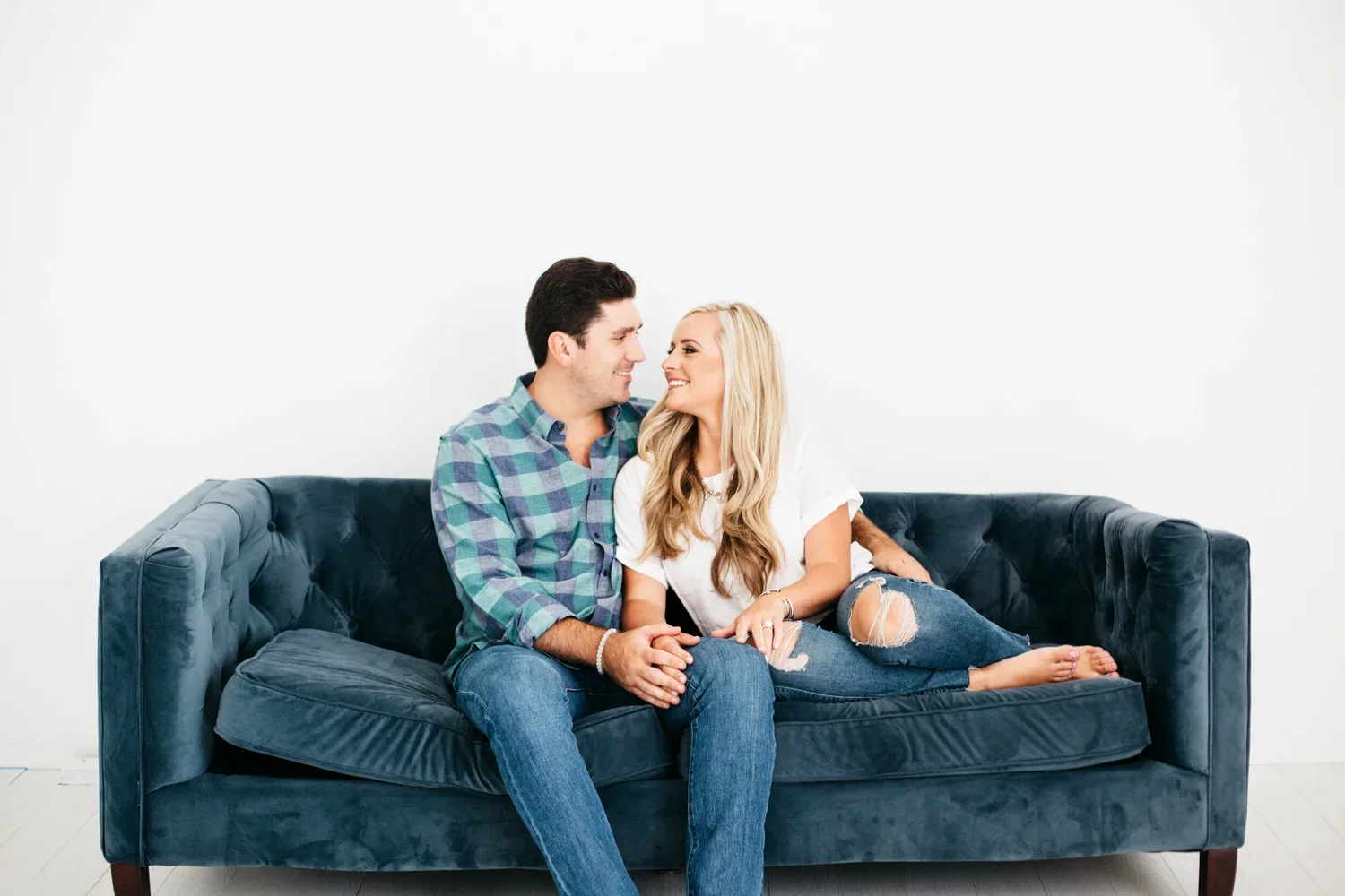 A smiling man and woman sitting closely on a dark blue velvet couch, facing each other and holding hands, in a room with a white wall background.