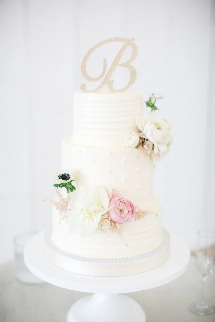 Two-tiered white wedding cake decorated with fresh flowers and a gold glittery letter 'B' cake topper.
