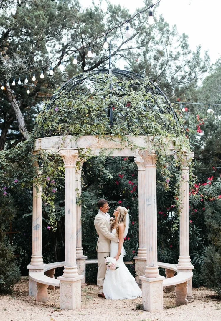 A bride and groom standing in an outdoor marble gazebo with columns, surrounded by lush green foliage and pink flowers, with string lights overhead.