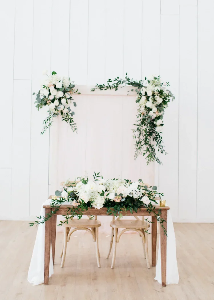 A floral wedding arch with white flowers and greenery, set against a white wooden wall, with a wooden table and two chairs decorated with flowers in front.