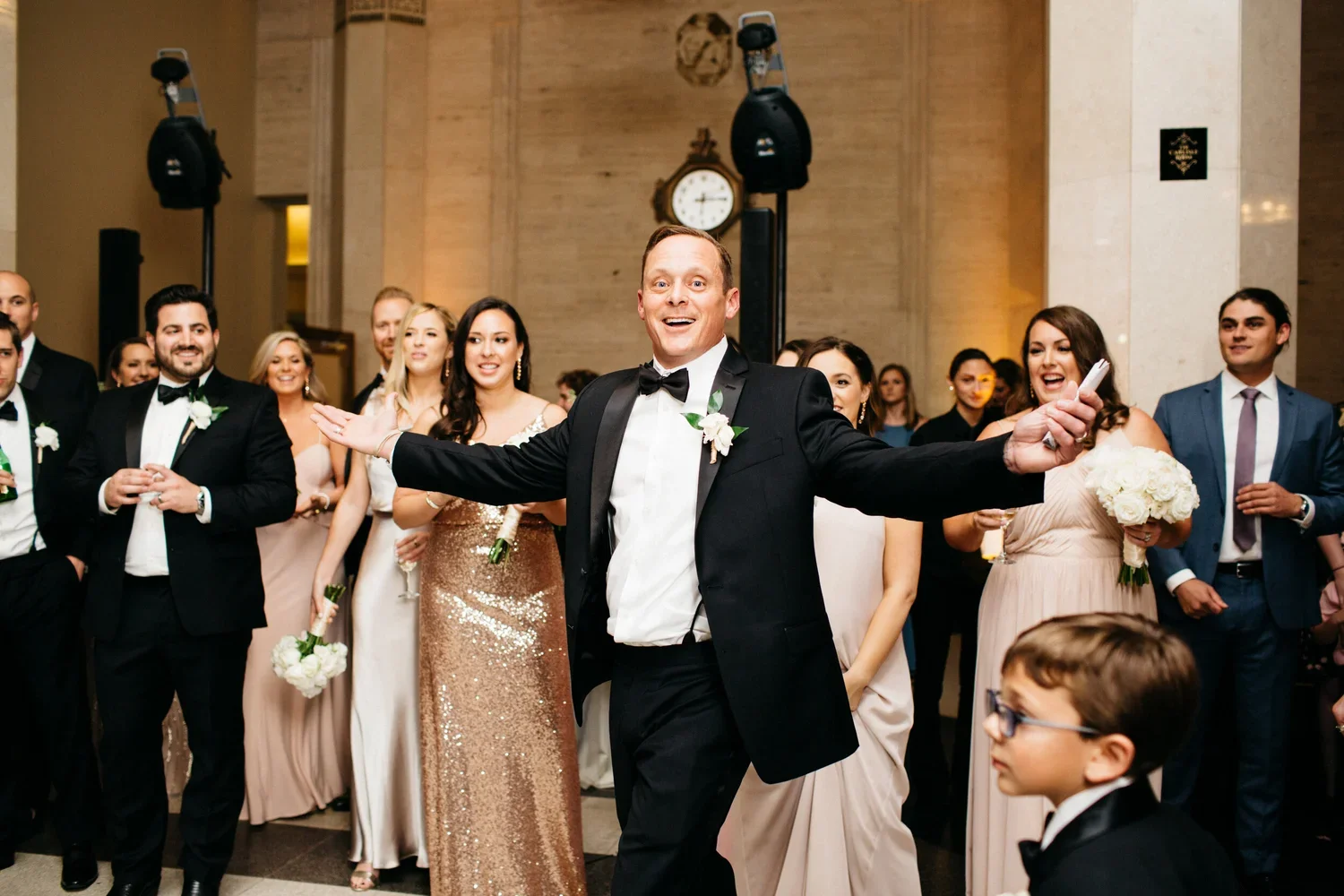 A groom in a tuxedo smiles with arms open at a wedding reception surrounded by guests in formal attire.