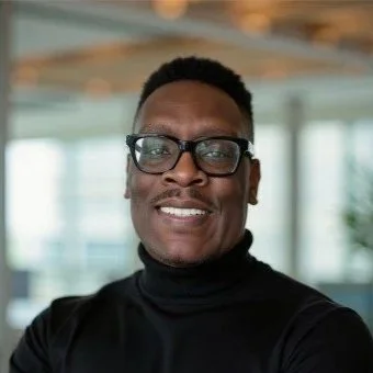 A man with glasses and short black hair smiling in an indoor setting with large windows.