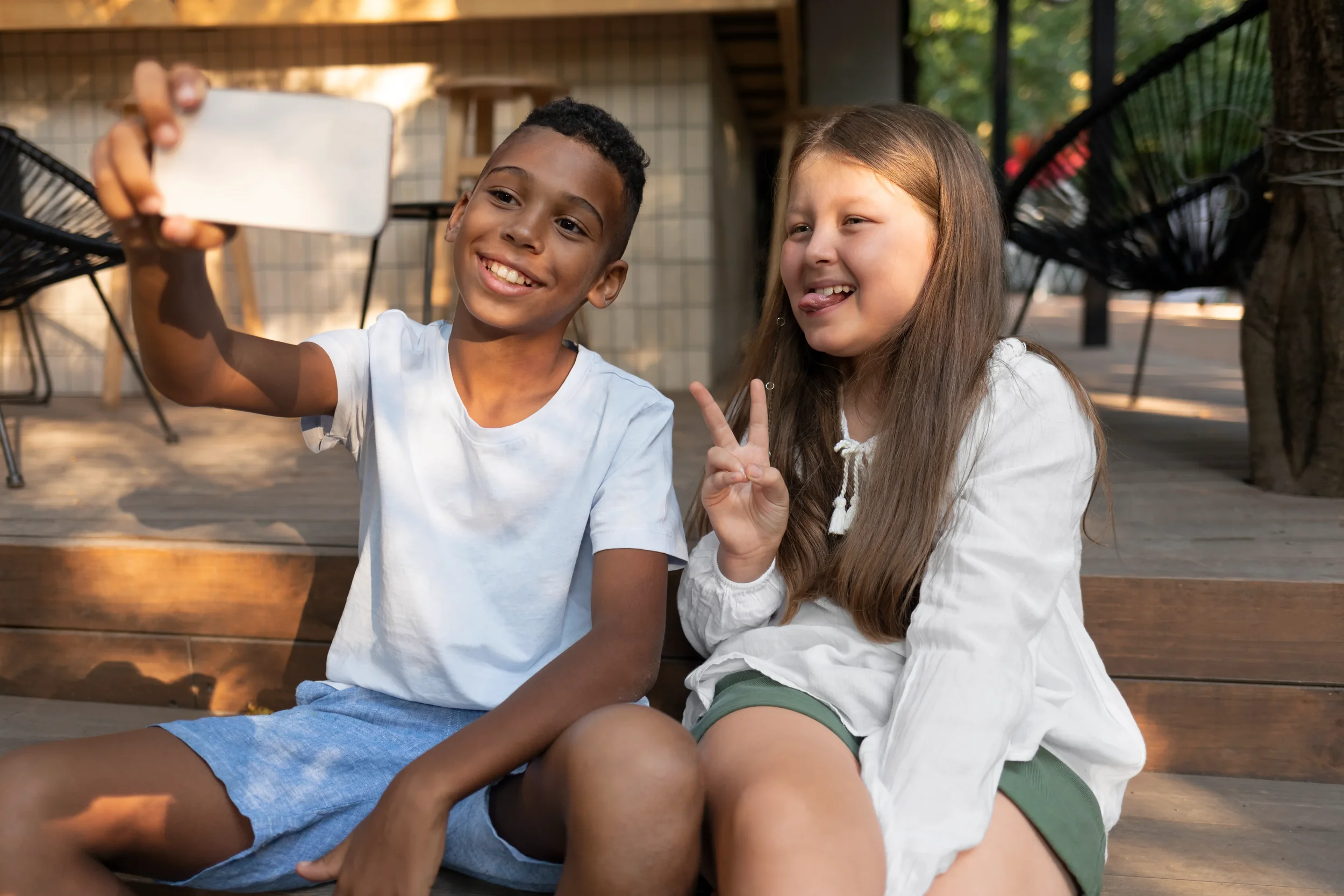 A boy and a girl sitting on a wooden porch taking a selfie. The boy is smiling and holding a smartphone, while the girl is making a peace sign with her fingers and sticking out her tongue. They are surrounded by outdoor furniture and greenery.