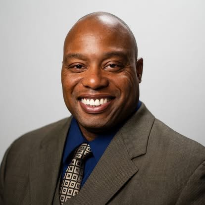 A smiling African American man in a gray suit, blue shirt, and patterned tie, posing against a light gray background.