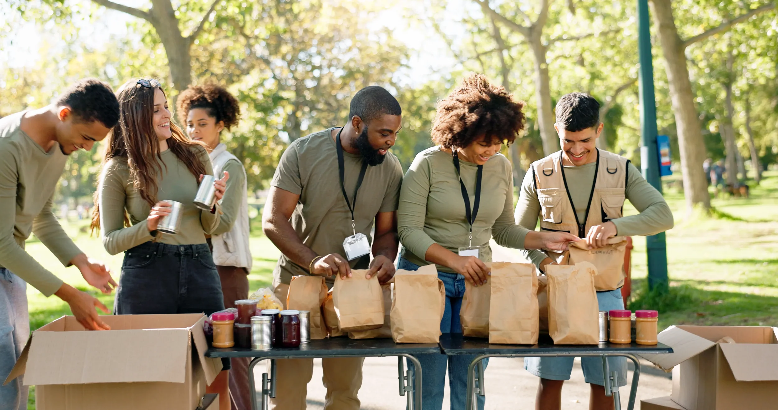 Group of people packing food into paper bags outdoors in a park on a sunny day.