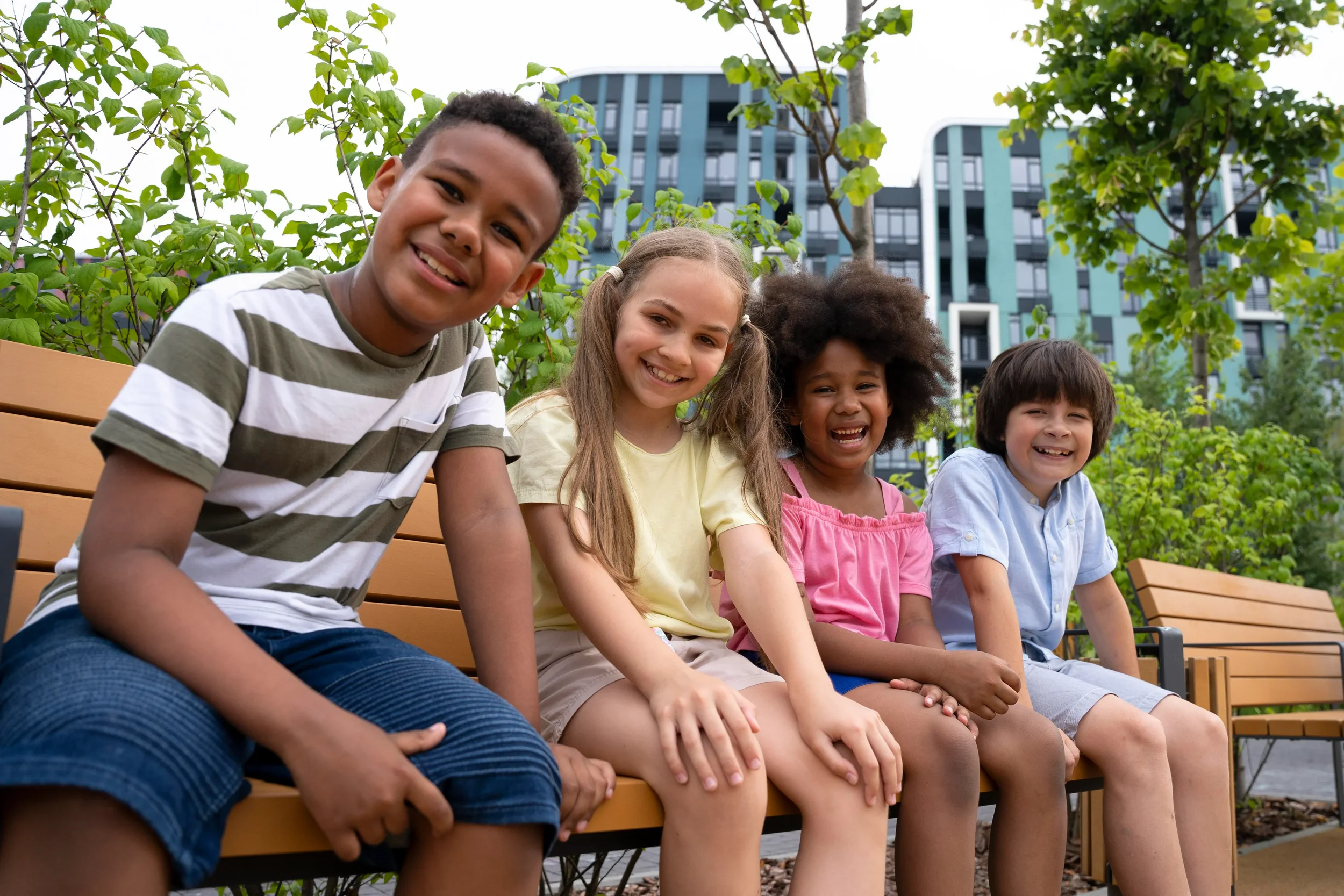 Four children sitting on a park bench smiling, with trees and modern buildings in the background.