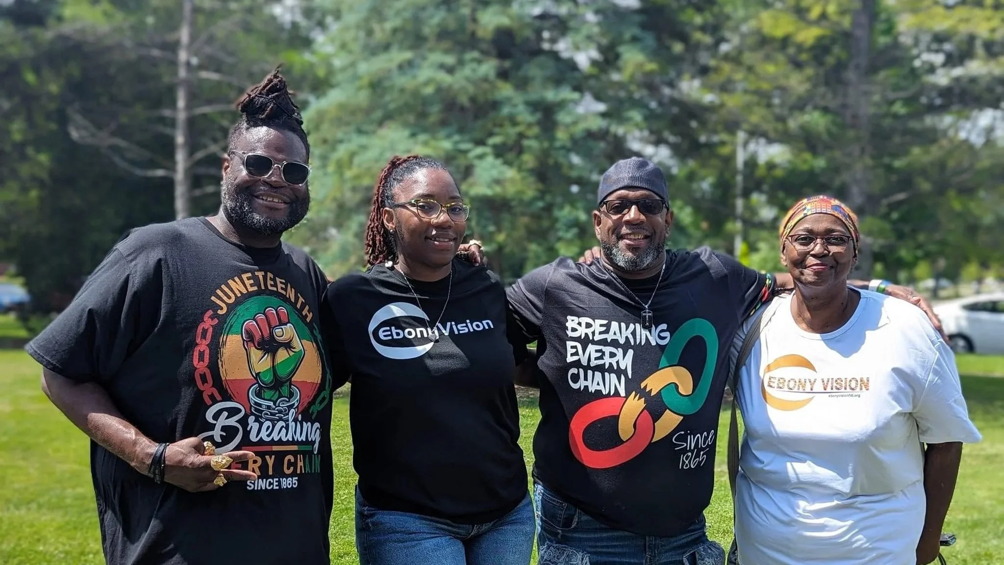 Four people standing outdoors on grass with trees in the background, smiling and wearing shirts with social justice messages and logos, including 'Breaking Chains' and 'Ebony Vision'.