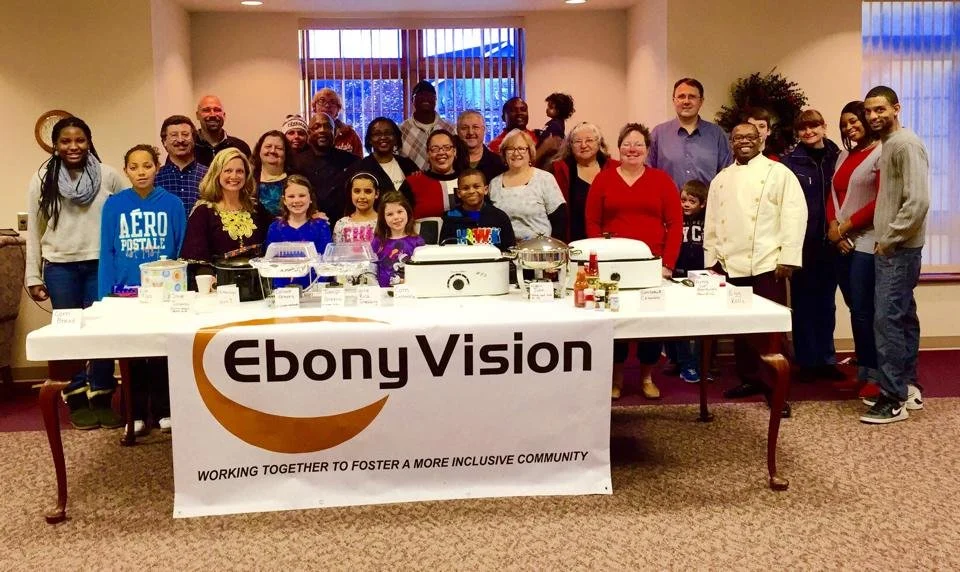A group of diverse people, including children and adults, standing behind a table with food and supplies, in a room with a banner that reads 'Ebonv Vision' and the slogan 'Working together to foster a more inclusive community.'