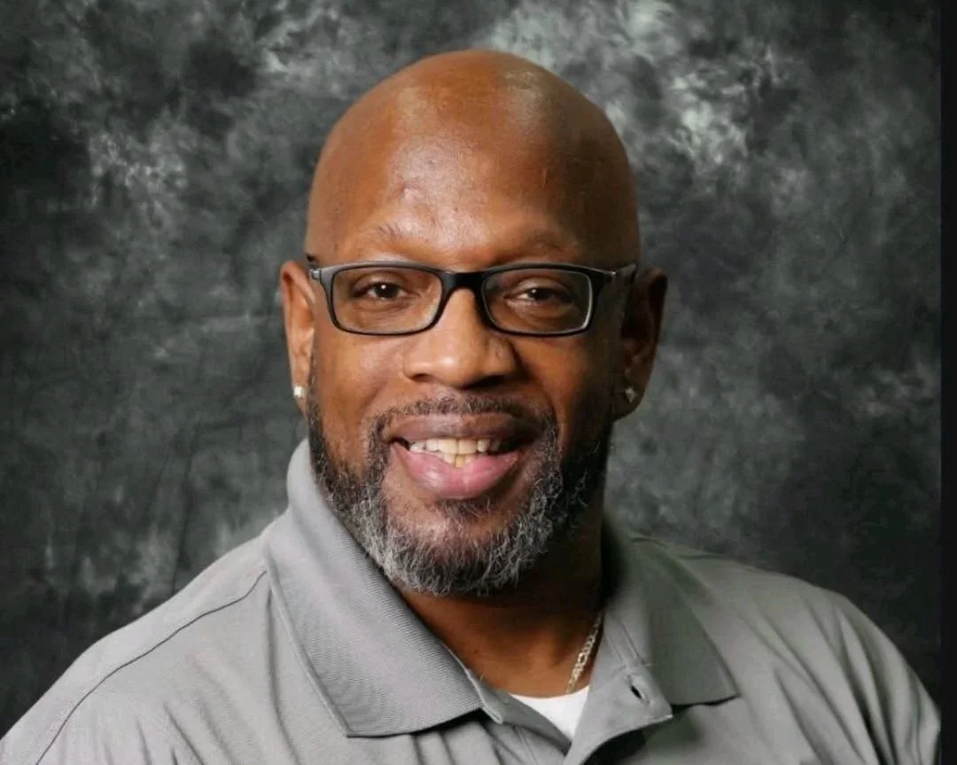 A middle-aged African American man with glasses, a beard, and earrings smiling in front of a dark, textured background.