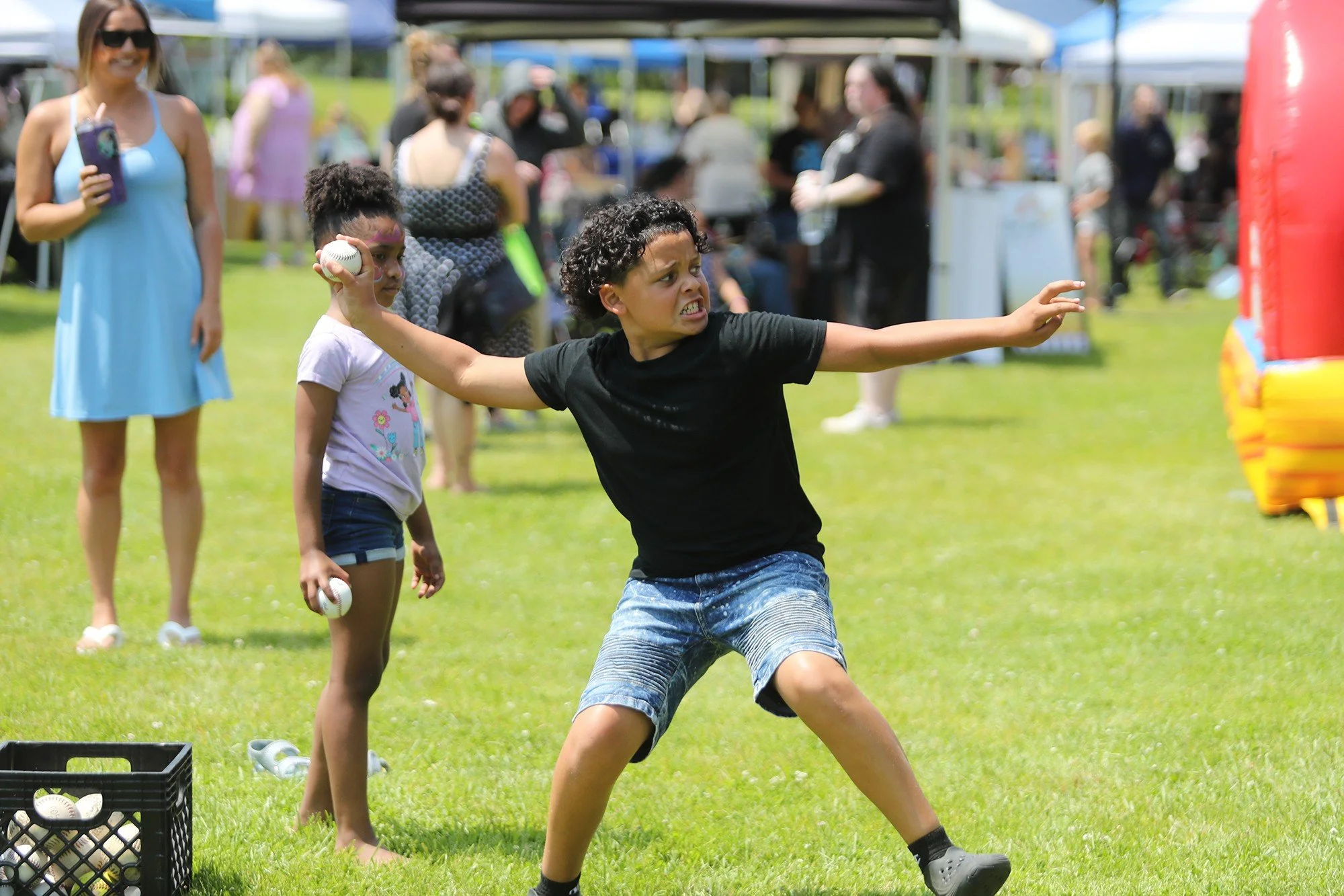 Children and adults at a park during a sunny day, with some children playing with balls and a woman smiling with a beverage