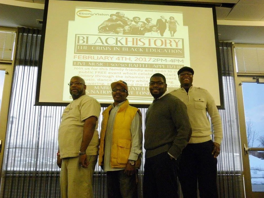 Four men standing in front of a large screen displaying a flyer about a Black History event, with sunlight visible through window blinds behind them.