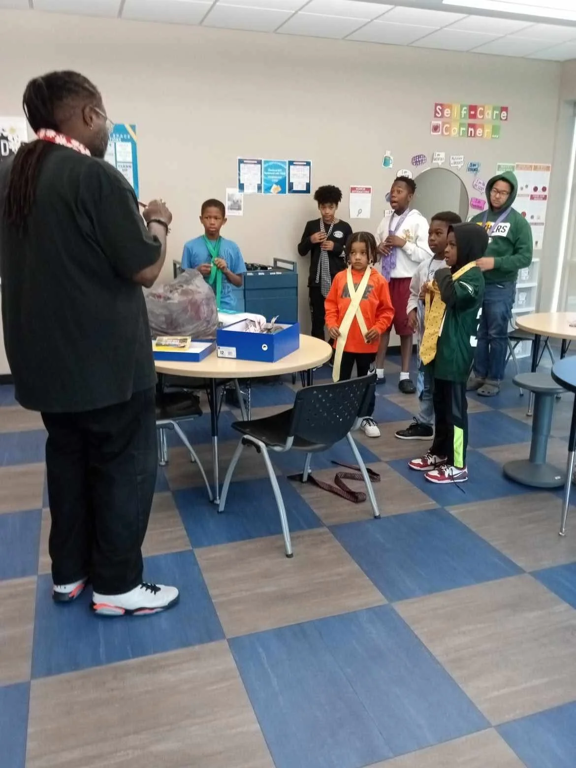 A group of children gathered around an adult in a room labeled as 'Self-Care Corner'. Some children are wearing special ribbons or sashes, and they appear to be engaged in an activity or discussion.