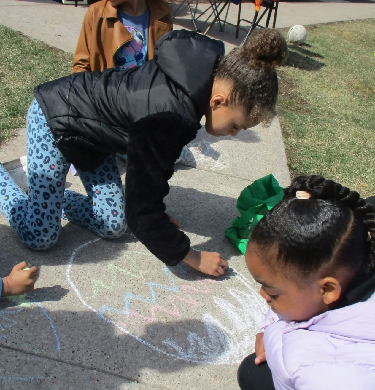 Two girls drawing with sidewalk chalk on a concrete sidewalk, one wearing leopard print pants and a black jacket, the other wearing a pink jacket and with braided hair, with a green gift bag nearby.